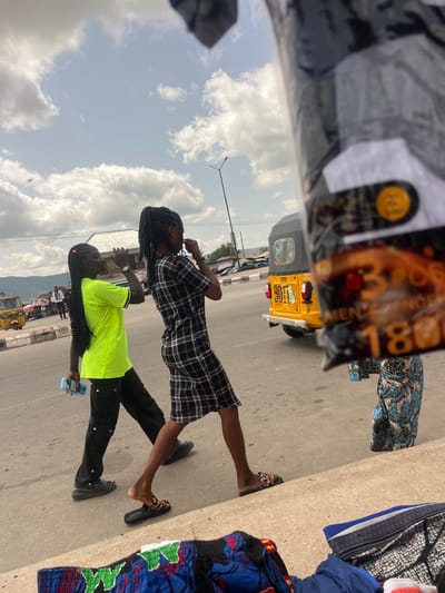 Two young women are standing on a street in Gulum, Nigeria, on a partly cloudy day. One woman is wearing a black and white plaid dress with cut-out shoulders, and her hair is styled in long, dark braids. She is looking up and to the side, seemingly lost in thought. The other woman is wearing a bright neon green t-shirt and black pants. Her hair is also in long, dark braids. She is holding a blue object, possibly a phone or a calculator, in her hand and looking downwards.
In the background, a yellow three-wheeled vehicle, a common mode of transport in Nigeria, is visible. Other vehicles and people can be seen further down the street, suggesting a bustling urban environment. A concrete barrier lines the side of the road, and piles of fabric, likely for sale, are displayed on the ground. The sky is filled with white, fluffy clouds, indicating fair weather. The image appears to be taken during daylight hours. The overall mood seems relaxed, with the women perhaps taking a break or conversing. No discernible text is visible in the image that provides specific context beyond what can be inferred from the scene.