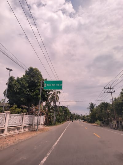 The image presents a daytime view of an asphalt road stretching into the distance, likely taken from a moving vehicle. The road features a white dashed line on the left shoulder and a double yellow line in the center, indicating two-way traffic.
On the left side of the road, a prominent green road sign is visible, displaying "Banda Aceh 178 km." Below the destination and distance, a smaller green shield shows the number "1" and "AH 25," likely indicating a route number and an Asian Highway designation.
Both sides of the road are lined with abundant, dense green vegetation, including large trees and palm trees, contributing to a lush, tropical setting. A decorative white fence with intricate patterns runs along the left side of the road, behind which a street light pole stands. Numerous utility poles and overhead power lines crisscross the upper portion of the frame against an overcast sky filled with scattered grey and white clouds.
In the distance, a motorcycle can be seen on the left side of the road, and a white car is further ahead, suggesting light traffic. The overall scene depicts a journey along a road in a semi-rural or suburban area. The time of day is daytime, under cloudy weather conditions. The location context indicates Ierhobglumpang, Indonesia, with the sign directing towards Banda Aceh.