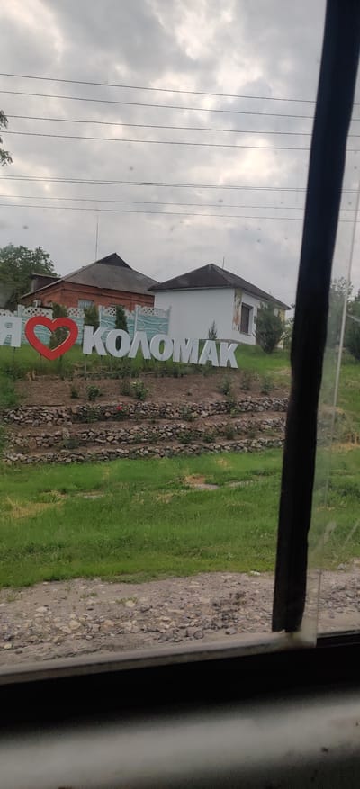 The image displays an outdoor scene, likely captured from a vehicle given the dark frame and visible smudges/reflections on what appears to be a window. The main focal point is a large, white monument sign set on the ground, prominently featuring the text "Я ❤️ КОЛОМАК" in Ukrainian Cyrillic. This translates to "I love Kolomak," with a vibrant red heart symbol replacing the word "love."
Behind the sign, two buildings are visible. The left building appears to be an older structure, possibly made of brick, with a dark, sloped roof. To its right, a light-colored, presumably white, building also has a dark, sloped roof and features decorative murals or paintings on its exterior walls, including a stylized window and a blue, wave-like pattern near its base.
The ground surrounding the sign and buildings consists of patches of green grass interspersed with areas of loose stones or rocks. The sky above is heavily overcast, suggesting a cloudy daytime. Several dark, thin power lines or utility wires traverse the upper part of the image against the gray sky. The scene is static, with no people or discernible activities taking place. The presence of the "I love Kolomak" sign acts as a clear location cue for Kolomak, Ukraine, conveying a sense of local pride.