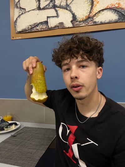 A young man with curly brown hair and a goatee holds a partially eaten pear in his right hand. He is wearing a black t-shirt with a white and red graphic design and a silver chain necklace. The man is looking towards the camera with his mouth slightly open. He is positioned in front of a blue wall, and a framed artwork hangs above him. To his left, a table is set with a placemat, a fork, a plate of food, and a glass of orange juice. The overall setting appears to be a domestic environment, possibly a dining room. The artwork on the wall bears a signature in the bottom left corner that reads "S. Greg-rio". While the image is not explicitly located in Somma Lombardo, Italy, the signature on the artwork suggests an artistic connection that may be relevant to that locale.