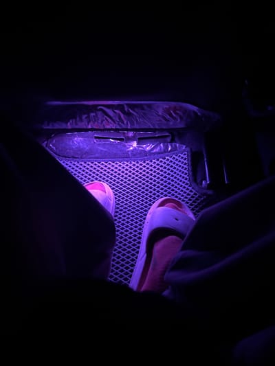 The image depicts the interior of a vehicle, likely a car, at night. Purple ambient lighting illuminates the scene, casting a mysterious glow on the footwell. In the foreground, two feet are visible, one wearing a pink sandal, and the other partially obscured by a dark fabric. The floor of the car is covered by a textured mat, possibly for protection or decoration, which is also bathed in the purple light. To the left, a dark, metallic object with a reflective surface is present, possibly a car pedal or part of the vehicle's chassis. The overall atmosphere is dimly lit and private, suggesting a moment of quiet or rest within the vehicle. The location is identified as Qarabalıq, Kazakhstan.