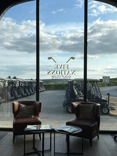 The image shows a view through a window, likely from a golf clubhouse in Méan, Belgium. The scene is set on a bright, cloudy day, possibly late morning or early afternoon.
The main subjects are golf carts parked in neat rows outside, viewed from an interior lounge area. Inside, two comfortable brown leather armchairs with cushions are visible, placed near small dark tables. One table has a magazine or brochure on it. The interior flooring appears to be made of wood planks.
Through the window, beyond the golf carts, there's a landscape with greenery and a sign visible in the distance. The sky is filled with dramatic, large white clouds against a light blue background.
Reflected on the window, there is text that reads "FIVE NATIONS GOLF CLUB" in gold lettering, with a golden golf club symbol to the left. The text is reversed due to the reflection. The overall impression is one of a modern, comfortable space offering a view of the golf course.
