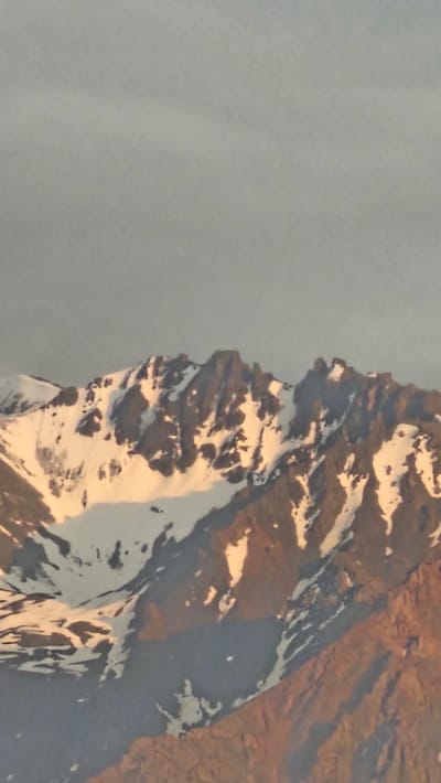 The image shows a mountain range with snow-capped peaks under a pale sky. The foreground is dominated by the rugged, rocky slopes of the mountains, with patches of snow clinging to the higher elevations. The lighting suggests it might be dawn or dusk, with a warm, golden hue cast on some of the rock faces. The overall scene is one of natural grandeur and remoteness. There are no people, animals, or man-made structures visible in the image. The location is specified as Панфилово, Kazakhstan, indicating a mountainous region within that country.