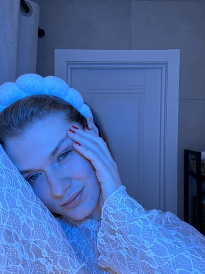 The image displays a close-up portrait of a person, likely female, lying down and looking directly at the camera. The image has a strong cool, bluish cast, suggesting low-light conditions or specific lighting choices.

The person has fair skin, blue eyes, and light-colored hair, partially visible around her face. She is wearing a white, lacy top or dress with long sleeves, the intricate floral or geometric lace pattern clearly visible, especially on the sleeve covering her right arm and shoulder. A soft, white, scalloped or bobble-textured headband is worn around her head, pushing back some of her hair. Her nails are painted a vibrant red.

Her pose is relaxed, with her head slightly propped up. Her right hand is resting gently on her forehead, fingers slightly splayed, with the red nail polish prominent. A subtle, serene smile plays on her lips, and her eyes are open, engaging directly with the viewer.

The background suggests an indoor setting. Above and behind her head, a white door frame or interior paneling is visible, featuring vertical grooves or sections within its design. To the left of the door frame (when the image is correctly oriented), a plain, light gray or blue-toned wall, possibly tiled, can be seen. In the lower left corner of the frame (when correctly oriented), there is a white, rolled-up towel or fabric item, and on it, a small, dark, circular object, possibly a hook or a decorative element. In the upper right background, a dark, rectangular object, potentially a shelf or picture frame, with some indistinct dark items on it, is visible.

The overall mood is calm and intimate, with the blue lighting contributing to a tranquil atmosphere. There is no visible text in the image.