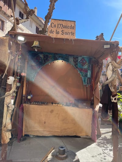 The image shows an outdoor booth or stall located in Hostalric, Spain. The booth has a sign that reads "La Moneda de la Suerte." It is decorated with various items including a bell, a light, animal skulls, horns, and fabrics. The backdrop appears to be decorated with a patterned textile in a turquoise color with an arched top. There is also what appears to be merchandise of some sort displayed for sale. The booth is situated in what seems to be a town square with buildings visible in the background. A rainbow-like light refraction is visible across the interior of the booth. The picture is taken during daylight under a clear blue sky.