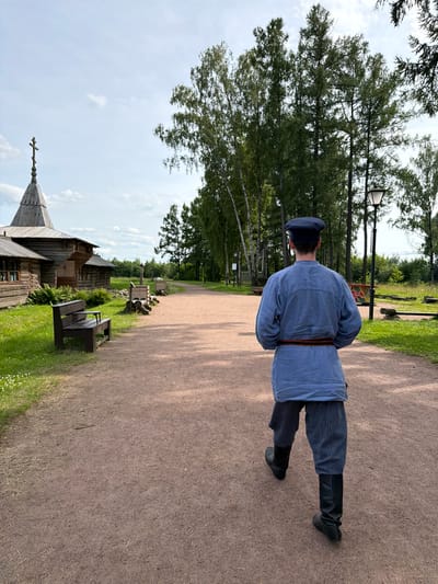 In Nevskii Parkleskhoz, Russia, a person walks away from the camera on a gravel path. They are wearing a blue, long-sleeved tunic, a striped pair of pants, black boots, and a dark cap. A decorative belt with red and black stripes is worn around their waist. To the left of the path, a traditional wooden church with a cross on its steeple stands next to a wooden bench. The background features a line of trees and a street lamp. The sky is overcast with scattered clouds.