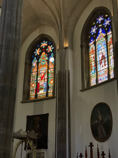 This media file captures the interior of a historic building, likely a church or cathedral, characterized by its architectural grandeur and religious art. The scene is dimly lit, with light primarily emanating from two large, ornate stained glass windows.
The upper section of the image features an arched stained glass window vibrant with blues, reds, greens, and yellows. It depicts religious figures, including a central figure with a halo, alongside other individuals. The upper parts of the arch are adorned with intricate floral or star-like patterns. To the right and slightly above this window, an oval-framed painting is visible on the light-colored wall. It portrays a solitary figure in flowing robes against a dark background, subtly highlighted by the ambient light. Further to the right, several dark, carved wooden spires or finials suggest decorative architectural elements.
A substantial dark beam or structural element spans horizontally across the middle of the image, dividing the upper and lower wall sections.
In the lower half, another large, arched stained glass window mirrors the style of the upper one, displaying vibrant religious narrative panels, with one figure prominently featured in a red cloak. To the right of this window, a rectangular painting is hung; it is very dark and indistinct, possibly depicting a multi-figured classical or religious scene. Below this painting, a highly ornate, gilded metal object with intricate scrollwork, possibly part of an altar or a large decorative fixture, reflects light.
The walls appear to be made of light-colored stone or plaster, providing a contrast to the darker architectural details and the glowing stained glass. The overall atmosphere conveys a sense of history, reverence, and artistic beauty. No specific activity or event is depicted, and no visible text is present. The setting is consistent with a religious building found in San Cristóbal De La Laguna, Spain.