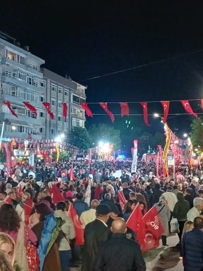 The scene is a large crowd of people gathered at night on a street in Silivri, Türkiye, located at coordinates 41.073 latitude, 28.244 longitude. The crowd is illuminated by streetlights and the glow of nearby buildings. The crowd is dense and packed together, with many people holding red flags with white crescents and stars, a symbol of the Turkish Republic. The flags are strung overhead, creating a canopy of red against the dark sky. The flags are a symbol of the Turkish Republic and likely represent the celebration of a national holiday or event. The buildings in the background are illuminated, casting a warm glow on the street. The crowd is diverse in age and gender. Some people are talking and gesturing, others are holding flags high in the air, and some are simply watching the scene unfold. The scene is one of celebration and unity. The crowd is gathered in front of a large building that appears to be a public structure, possibly a government building or community center. The building is illuminated with warm lights, and there is a large sign above the entrance that reads "CHP," which is the abbreviation for the Republican People's Party, a major political party in Turkey. The scene is likely a political rally or a celebration of a political event. The presence of flags and banners suggests a political gathering, and the presence of the CHP sign further indicates that the event is associated with the party.