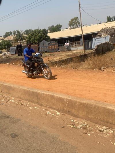 An adult male is observed operating a black motorcycle on an unpaved, reddish-brown dirt road in Lapai, Nigeria. The individual wears a blue short-sleeved collared shirt and dark trousers. The motorcycle features silver spoked wheels and a chrome exhaust. In the foreground, a concrete curb separates the dirt road from a darker, paved surface, scattered with wood shavings and debris. The background reveals a commercial establishment displaying multiple finished metal gates of various designs, backed by a large building with a light-colored corrugated metal roof. Dry, light brown vegetation covers parts of the ground, interspersed with green trees and a palm tree under a clear, bright sky. A wooden utility pole with overhead wires is visible on the right side of the dirt road.