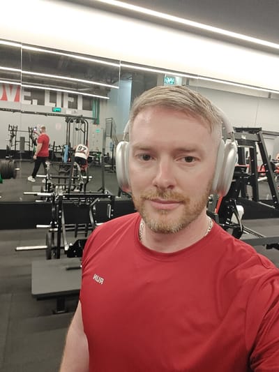 A light-skinned male with short blonde hair and a reddish-blonde beard takes a selfie in an indoor fitness facility, located in Nahariya, Israel. He wears a red short-sleeved athletic shirt with the mirrored text "RUN" visible on the chest, a silver chain, and silver over-ear headphones. His gaze is directed towards the camera.

The background consists of a large mirrored wall reflecting the gym's interior. In the reflection, another male individual wearing a red shirt and dark pants is visible, holding a mobile phone, positioned near a weight rack with barbells. Further back, a second male individual wearing a white and black numbered tank top (number "97" is legible) and dark shorts is seated on an exercise machine. The gym features various weightlifting equipment, including black-framed racks, barbells with black plates, and benches. Overhead, bright white strip lighting illuminates the space. Reflected signage on the wall includes partially obscured English text, possibly "LOVE.LIFT," and visible Hebrew text "צאו" (Exit), indicating an Israeli location. The floor is covered with dark gray or black matting.