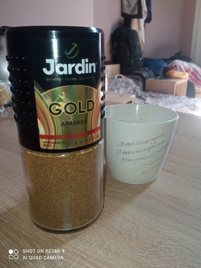 A close-up, eye-level, medium shot shows a jar of Jardin Gold Arabika instant coffee and a white ceramic mug on a light brown wooden table. The coffee jar is black with a gold and green label that reads "Jardin" and "GOLD ARABIKA". The mug is to the right of the coffee jar and has handwritten text in Russian that appears to be a personal note. The background is out of focus, showing a wall and some furniture. The lighting suggests an indoor environment, possibly a home or office. The image is watermarked with "SHOT ON REDMI 9 AI QUAD CAMERA" in the bottom left corner. There are no people visible in the image.