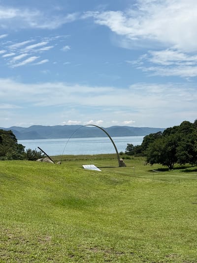 An outdoor landscape scene on the island of Naoshima, Japan, showcases a serene natural environment with a prominent art installation. The foreground is a lush green grassy hill sloping gently towards a body of water. In the mid-ground, a large, curved metallic sculpture dominates the scene, arching gracefully over the landscape. To the left of the sculpture, a large, rough-hewn boulder is visible, with a dark, angled metal pole leaning against it. A white, flat object, possibly part of another installation or signage, rests on the grass in front of the curved sculpture.
The background features a tranquil blue body of water that stretches across the horizon, meeting a chain of distant, hazy blue mountains. The sky above is a vibrant blue, dotted with fluffy white clouds. Dense green trees line the edges of the scene, framing the water and mountains. The overall impression is one of art integrated harmoniously with nature, typical of the artistic landscape found on Naoshima.