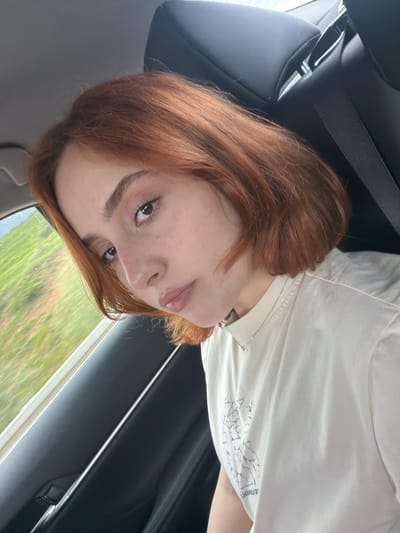 A young woman with auburn hair is in the backseat of a car, wearing a light-colored t-shirt with a graphic design. She is looking at the camera. The background shows the car's interior, including the headrest, seatbelt, and door panel. Outside the window, there is blurred greenery. The exact location within Tserovani, Georgia, cannot be determined from this image.