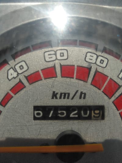 The image presents a detailed, static, close-up view of a vehicle's instrument cluster, contextualized as being within La Roche-en-Ardenne, Belgium. The primary subject is a grey-textured speedometer dial displaying speed increments of "40", "60", and "80" km/h, with "km/h" units printed below. Red rectangular segments mark intervals on the speed scale. Centrally positioned below the speed dial is a black rectangular digital odometer, which registers "075209" units. At the bottom of the frame, a segment of a horizontal orange bar, possibly an indicator, is visible, partially obscured by a circular black button or cap. The instrument cluster surface shows numerous scratches and minor particulate matter. A strong light source, consistent with direct sunlight, creates a significant lens flare and glare in the upper central area of the image, affecting visibility of the upper dial segment. No people, active interactions, or additional environmental details beyond the immediate instrument panel are discernible.