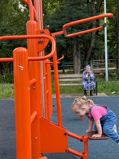 In Viazniky, Russia, on an outdoor fitness equipment area, a young Caucasian girl with blonde pigtails and a pink shirt and denim overalls is crouched on an orange exercise machine. Her hands are on the machine's seat, and she appears to be climbing onto it. In the background, an elderly Caucasian woman with white hair is seated on a wooden bench, holding a cane and wearing a brightly patterned robe and headscarf. The fitness equipment is made of orange metal tubes. Behind the bench, trees and some fencing are visible, suggesting a park or public space. The ground is covered with blue rubber matting.