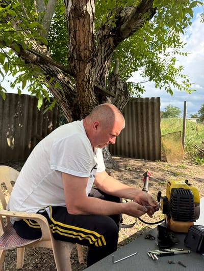 A man in a white t-shirt and black athletic pants with yellow stripes is sitting on a plastic chair and working on a yellow and black gas-powered string trimmer. He is holding a hex wrench and appears to be tightening or loosening a bolt on the trimmer's engine. Various tools, including other hex wrenches and bolts, are laid out on a gray table in front of him. The scene is outdoors, with a large tree providing shade. In the background, there is a corrugated metal fence, some dry vegetation, and a cloudy sky. The ground is covered in dirt and small stones. The man is fair-skinned with a shaved head and is looking down at his work. The overall impression is of a casual outdoor repair or maintenance activity.