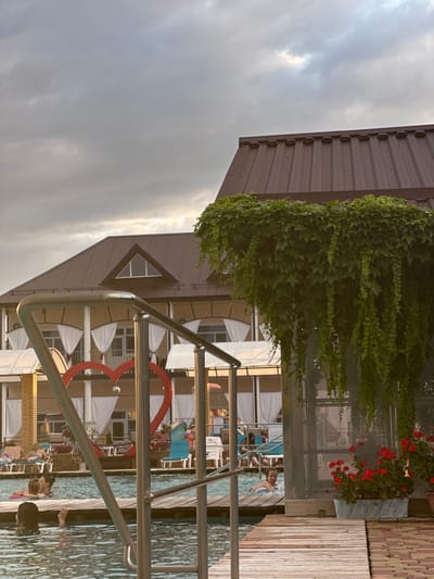 The image captures an outdoor swimming pool area, likely a resort or spa, under an overcast sky. In the foreground, a metal railing of a staircase leads into the water, with a wooden deck bordering the pool. Several people are in the pool, some swimming and others relaxing. One group in the water appears to be engaged in conversation. In the mid-ground, a large red heart sculpture is visible. Behind the pool, a building with a brown metal roof and a yellow facade is visible, adorned with white curtains and awnings. Lush green ivy hangs from an overhang of the building on the right side, and potted red flowers are also present. The scene suggests a leisure activity in a warm climate. While no specific landmarks clearly identify it as Yaroslavskaya, Russia, the setting is consistent with recreational facilities found in such a region.