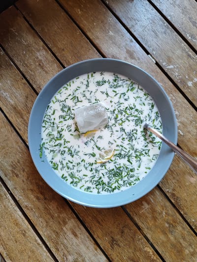 A close-up overhead shot shows a light blue bowl of cold soup, likely okroshka, filled with a white creamy liquid, chopped green herbs such as dill and chives, and small pieces of cucumber. A large ice cube sits in the center of the soup, accompanied by a thin slice of lemon. A silver spoon rests on the right edge of the bowl, its handle extending out of frame. The soup is placed on a rustic wooden table with prominent grain and a weathered appearance. The background is entirely composed of this wooden surface, suggesting an outdoor or casual indoor setting. The lighting is bright, creating soft shadows and highlighting the textures of both the food and the table. There is no visible text in the image. The scene conveys a sense of refreshment and simple dining, possibly during a warm day in El Campello, Spain.
