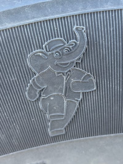 The image shows a close-up of the textured sole of a shoe, featuring an embossed cartoon elephant wearing a hat and a collared shirt. The elephant is depicted in profile, with its trunk raised and a smiling expression. The sole has a ribbed pattern running vertically. The scene is set on a black, textured surface, likely the bottom of footwear. No specific location in Zarech'e, Russia, can be identified from the image.