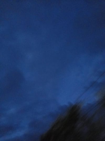 The image depicts a dark, blurry scene with a dominant deep blue hue, suggesting twilight or nighttime. The sky occupies most of the frame, with subtle variations in darkness that could indicate clouds or atmospheric haze. In the lower right portion of the image, there is a dark, out-of-focus mass that appears to be foliage or trees, possibly silhouetted against the sky. Faint, diagonal lines are visible, which could be power lines or some other distant structures. The overall impression is one of a dimly lit, possibly windy, outdoor environment. The location context of Khwap, Abkhazia, points to a natural setting, potentially a rural or semi-rural area. The lack of sharp detail and the muted colors evoke a sense of mystery or quietude. No people, text, or specific events are discernible due to the blurriness and low light.