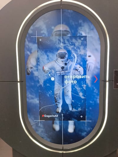 This media file shows a digital display featuring a stylized image of an astronaut against a backdrop of Earth viewed from space. The astronaut is in a full white spacesuit, helmet with visor, and appears to be floating. There is a superimposed image or graphic of the astronaut, with some lines and circles suggesting augmented reality elements.
On the screen, in large white Cyrillic text, are the words "отправить фото", which translates to "send photo". To the right of this text, there is a red arrow pointing to the right. Below the astronaut's legs, the hashtag "#GagarinAR" is displayed in red text. The background is a vibrant blue, suggesting the Earth's atmosphere and oceans as seen from orbit.
The display itself is framed within an oval-shaped, dark grey structure with a bright white light strip around its inner edge. The image on the screen appears to be a photograph or a rendered graphic, potentially interactive. The setting is likely an exhibit or a public space, given the presentation style. The overall impression is one of a space-themed interactive experience, possibly a photo booth or an AR application related to Yuri Gagarin. The context of "Vjazovka, Russia" suggests a local event or exhibition in that area. There are no clear indicators of time of day or weather.