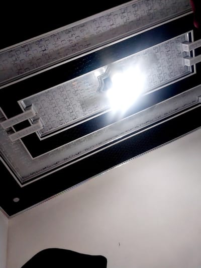 The image shows a modern, illuminated ceiling with a geometric pattern. The ceiling features a black background with a grid of translucent panels overlaid with a repeating pattern of squares and rectangles. Recessed lighting illuminates the panels from behind, creating a bright, diffused glow. Some areas of the ceiling are framed with silver trim. The overall impression is one of contemporary design and sophisticated lighting. The image was taken in Jeppee, Indonesia, a city known for its modern architecture and urban development.