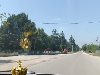 The image shows a road in Hiliuți, Moldova, with a large decorative pitcher on the left side and an "I LOVE HILIUȚI" sign. The sign has a red heart and white lettering. The road is paved and appears to be a rural or suburban area. Trees line both sides of the road, and there are power lines overhead. The sky is clear and blue. In the foreground, a golden crown with a cross is visible on a dashboard, suggesting the photo was taken from inside a vehicle. The overall scene depicts a scenic view of Hiliuți, likely a welcome sign for visitors.