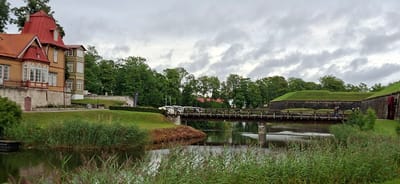 The image depicts a picturesque scene in Kuressaare, Estonia, likely during daylight under an overcast sky. The composition is dominated by a canal or moat with lush green reeds lining its banks. A wooden bridge spans the water, with a few people visible crossing it. Beyond the bridge, there's a well-maintained grassy area and a section of a stone wall, suggesting a historical fortification.
On the right side of the image, a charming old building with a distinct yellow facade and red tiled roof is visible. The building features decorative window frames and balconies, indicative of historical architecture. Trees with dense green foliage are scattered throughout the scene, adding to the natural beauty. The overall atmosphere appears peaceful and serene, characteristic of a well-preserved historical site. No visible text is present in the image.