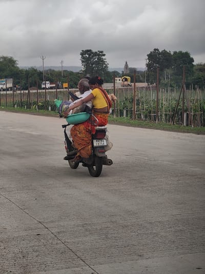 In Walsawangi, India, on a paved road under an overcast sky, an elderly man and a younger woman are riding on a motorcycle. The man, with white hair and a beard, is driving, while the woman, wearing a yellow top and an orange and red patterned sari, is seated behind him, holding a green bowl. Both are facing away from the camera. The motorcycle is loaded with various items, including bags and what appears to be a black umbrella strapped to the back. In the background, there are trees, a fence, a temple structure with a spire, and some vehicles on a distant road. The road surface shows signs of wear and tear.