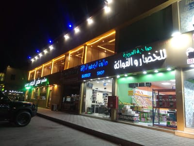 The image depicts a street scene at night in Al Khobar, Saudi Arabia. A black SUV is parked on the street in front of a row of businesses. From left to right, these include a restaurant named " مطعم سفير الجنوب محمية", a barbershop named " صالون الجدهور الانيقة BARBER SHOP ", and a produce shop named " صاحية العزيزية للخضار والفواكة مؤسسة". The produce shop appears to have a wide variety of fruits and vegetables on display. The facades of the shops are well-lit, and there are streetlights overhead. The sidewalk is paved with interlocking gray pavers.