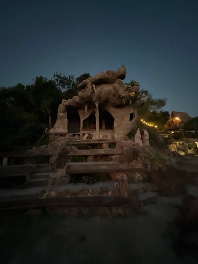 The image depicts a dark, twilight scene in Karimunjawa, Indonesia, featuring a unique, cave-like structure with faux rock formations and a rustic wooden cabin in the background. The cave-like structure, constructed from what appears to be concrete molded to resemble rock, has several openings and is illuminated by subtle lighting, highlighting icicle-like formations hanging from its overhang. A stone staircase with wooden railings leads up to the structure, with some small pink flowers visible in planters. To the right, a traditional Indonesian wooden cabin on stilts is visible, adorned with warm, string lights that create a festive atmosphere. The surrounding environment is lush with trees and foliage, silhouetted against the darkening sky. The overall impression is of a whimsical, possibly themed, establishment set in a tropical location.