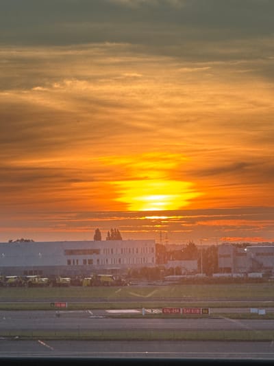 This is an image of a sunrise over an industrial area near Zaventem, Belgium. The sky is filled with warm hues of orange and yellow, with streaks of gray clouds. In the middle ground, there are several large, modern industrial buildings. In front of the buildings, a row of yellow construction vehicles is parked. The foreground shows a section of an airport tarmac with runway markings. The overall scene suggests an early morning at an airport or industrial park.