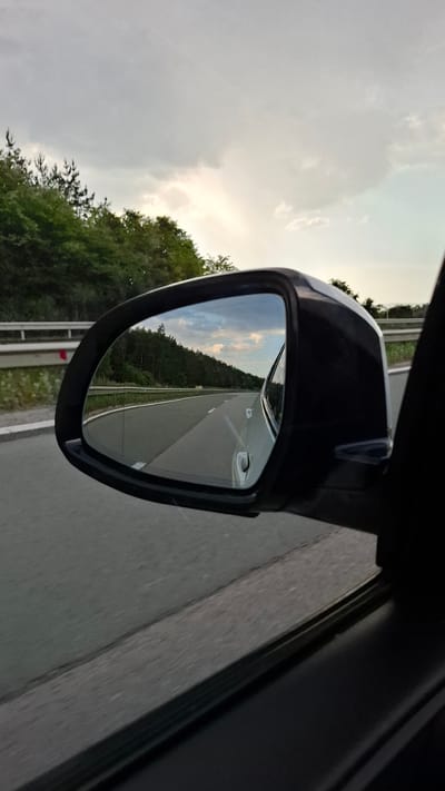 A car's side mirror reflects a highway cutting through a wooded area under a cloudy sky. The mirror, mounted on a black housing, captures the road stretching into the distance, lined by a guardrail and dense green trees. The sky in the reflection shows patches of lighter clouds, possibly indicating a sunset or sunrise. The foreground shows a portion of the car's window and interior. The scene appears to be captured from a moving vehicle, as evidenced by the motion blur on the road outside the mirror. The location is identified as Gorna Vasilitsa, Bulgaria.