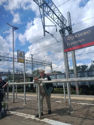 The image shows a station platform in Fryazevo, Russia, under a cloudy blue sky. A sign above the platform reads "Фрязево Fryazevo". Two women are present: one in the foreground leans on a white railing, looking to her left, while another woman stands further back on the left, partially visible. A bouquet of white flowers in a grey bag is on the ground near the woman in the foreground. The platform is paved with brick, and a train is visible in the background, composed of several grey freight cars. Overhead, there are power lines and structural supports for the railway. A yellow triangular sign with a train symbol and Russian text is mounted on a pole to the left of the foreground woman.