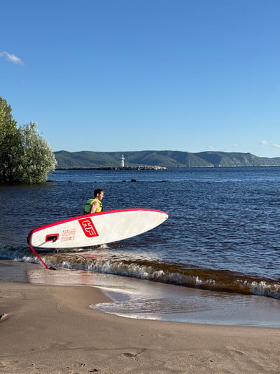 This media file depicts a man in a yellow life jacket standing next to a stand-up paddleboard on a sandy beach. The paddleboard is white with red trim and the "HF" logo is visible on its side. The man is looking out at a large body of water, likely a river or lake, with rolling hills and trees in the background. A lighthouse is visible in the distance. The sky is clear and blue, suggesting a sunny day. The scene appears to be set in the afternoon. The overall mood is peaceful and recreational, with the man appearing relaxed and contemplative as he prepares to engage in or has just finished a water activity. The location is identified as Volzhskiy Utes, Russia. The text on the paddleboard, besides the "HF" logo, includes some illegible instructions or branding.