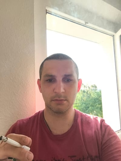 This is a medium shot, selfie-style photograph of a man in his late 20s to early 30s. He has short, dark hair, a light beard, and is looking directly at the camera with a neutral expression. The man is wearing a dark pink t-shirt. His right hand is in the foreground, holding a lit cigarette with smoke rising from it.

The setting appears to be indoors, near a window. To the left of the man is a textured, cream-colored wall. To his right, the window reveals a bright, sunny outdoor scene with lush green trees. The light coming from the window is very bright, suggesting it is daytime, likely mid-morning or afternoon, with clear weather. There are faint wisps of smoke visible near the top of the window frame.

There is no visible text in the image. The overall mood is casual and relaxed. The location context is Wolgast, Germany.