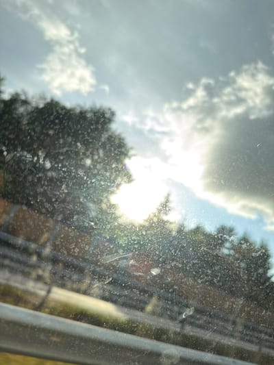 The image depicts a view through a dirty window, likely of a vehicle, looking out at a sunlit sky with clouds and trees. The sun is partially obscured by clouds and trees, creating lens flare and a bright glow. The foreground is dominated by the dirty window surface, which is covered in numerous spots and streaks. In the middle ground, blurry green trees line a road or pathway. The background consists of a bright sky with scattered white and grey clouds. The scene appears to be captured outdoors during the day. There are no people or specific landmarks that would indicate the location within Fjärås, Sweden, beyond the general outdoor environment.