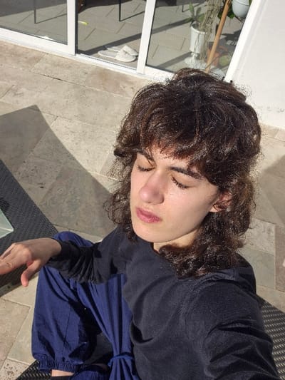 The scene depicts an individual seated outdoors on a light-colored tiled patio or balcony, presumed to be within a private residential property in Châtelaillon-Plage, France. The person, with dark, curly hair, is shown in a high-angle view, eyes closed and face tilted upwards, indicating exposure to strong, direct sunlight. They wear a dark long-sleeved shirt and loose blue pants. A dark, woven outdoor furniture surface is partially visible beneath their right hand. In the background, a white-framed glass sliding door reflects an interior space containing two white slippers on the floor and a potted plant. Distinct shadows are cast across the paved surface by the bright illumination.