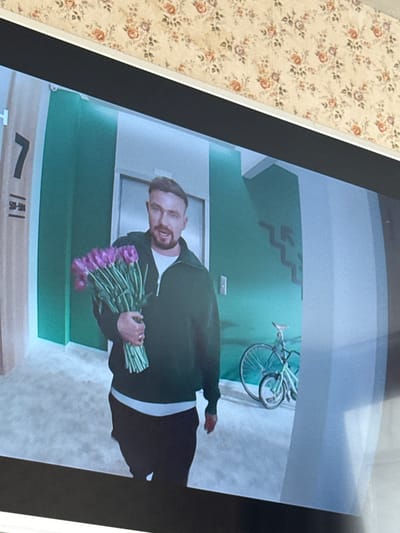 The image shows a man holding a bouquet of purple tulips. He is standing in a hallway with green walls. There is a door behind him. A bicycle is mounted on the wall in the background. The location appears to be an apartment building, possibly in Russia, given the city Nevzorovo is specified.