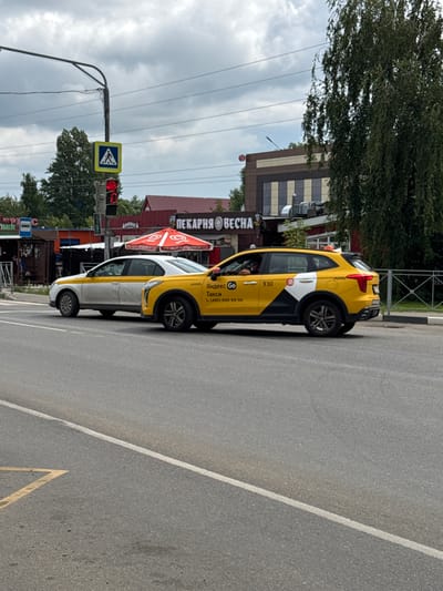 A street scene in Rastunovo, Russia, features two yellow and white Yandex taxis, one in the foreground and one behind. A traffic light is red, and a pedestrian crossing sign is visible. In the background, a building with "PEKARNYA VESNA" signage is present, along with trees and a cloudy sky. The road has painted lane markers.