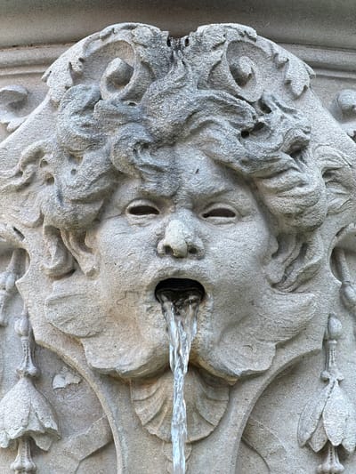 The image shows a stone fountain in Grad Opatija, Croatia. The fountain features a sculpted face with a hole in its mouth, through which water flows. The face is weathered and textured, with stylized hair and facial features. The water is clear and creates a smooth, linear stream. The stone surface is light gray and appears aged.