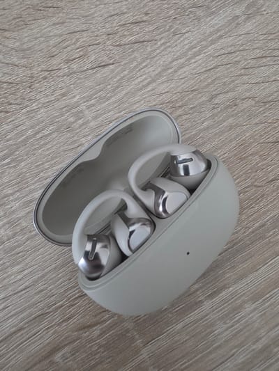 The image shows a pair of white and silver wireless earbuds in their charging case. The case is light gray and slightly open, revealing the earbuds nestled inside. The earbuds have a sleek, modern design with a silver metallic finish on the ear tips and a white ear hook. The charging case is placed on a light-colored, textured surface that resembles wood grain. There is no indication of the city of Oberhausen, Germany, or any environmental or human elements.