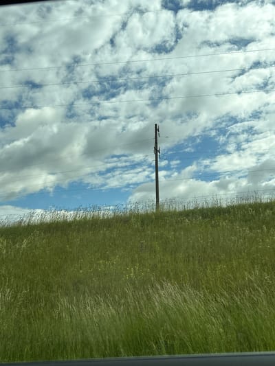 This is a photograph taken from a vehicle, looking out towards a grassy hillside under a cloudy sky. A single wooden utility pole stands on the right side of the frame, with several power lines running horizontally across the sky. The hillside is covered in dense, green grass, with some taller, wispy blades of grass and a few scattered yellow wildflowers. The sky is a mix of blue patches and large, white, puffy clouds. The overall impression is a rural or roadside scene. The location is identified as Malevichi Rural Council, Belarus.