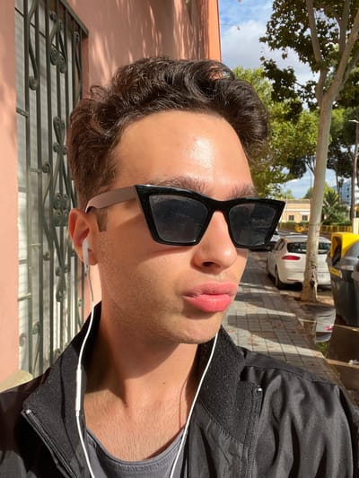 The image displays a close-up portrait of an individual, likely male, with dark, wavy hair, wearing black cat-eye sunglasses and white wired earphones. The individual's face is illuminated by direct sunlight, casting shadows, and features slightly pursed, pink-toned lips. The person wears a black zipped jacket over a grey t-shirt. The scene is located outdoors in Picanya, Spain. In the immediate background to the left, a portion of a salmon-pink building wall with dark, decorative metal security bars is visible. To the right and receding into the mid-ground, a concrete sidewalk is present, featuring tree shadows and a small puddle. Several parked vehicles, predominantly white sedans (one identifiable as a SEAT), line the street alongside the sidewalk. Two waste bins, one yellow and one dark grey, stand on the sidewalk. Green trees and a partially cloudy blue sky are visible in the upper right background.
