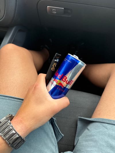 A person is holding a can of Red Bull and a black Icc Vape in what appears to be the interior of a car. The person is wearing a watch on their left wrist. The location is not evident from the image. No part of Hadzhidimitrovo, Bulgaria can be identified.