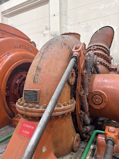 The image displays a detailed view of a large-scale industrial turbine and generator assembly, located within a hydroelectric power plant in Pellafol, France. The primary subject is a reddish-orange colored Francis turbine, manufactured by "ATELIERS NEYRET-BEYLIER & PICCARD-PICTET" in Grenoble in 1913, rated at 2000 CV, as indicated on a black metallic plaque affixed to its structure. This turbine connects to an "ALSTHOM BELFORT" generator, partially visible on the left.

The machinery exhibits significant wear, corrosion, and peeling paint, suggesting age and prolonged operation or disuse. A prominent, thick, grey metal rod extends diagonally across the mid-ground, connected to the turbine structure via an orange clevis. To the right of this rod, a series of rusty coiled springs and linkages are integrated into the turbine's mechanism. A red warning sign with white text reading "ATTENTION FONTE" (Caution: Cast Iron) is affixed to the lower section of the turbine. A green pipe is visible along the bottom edge, connecting to other unseen components.

The immediate environment consists of worn, light-colored walls with peeling paint and discoloration, typical of an industrial facility. Portions of a metal grate floor are visible on the left, with an unidentifiable floor surface to the right. No individuals are present in the scene.