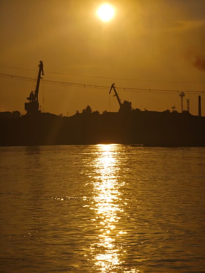 This is a photograph of a sunset over a body of water, likely a river, in the city of Lesosibirsk, Russia. The sun, a bright orb in the upper center of the frame, casts a warm, golden light across the scene. The water in the foreground reflects the sunlight, creating a shimmering path of light that stretches across the surface. In the background, silhouetted against the bright sky, are several industrial cranes and structures, suggesting a port or industrial area. Trees and other vegetation are visible along the distant shore, forming a dark line against the horizon. The overall atmosphere is serene and majestic, capturing the beauty of a natural phenomenon in an industrial setting.