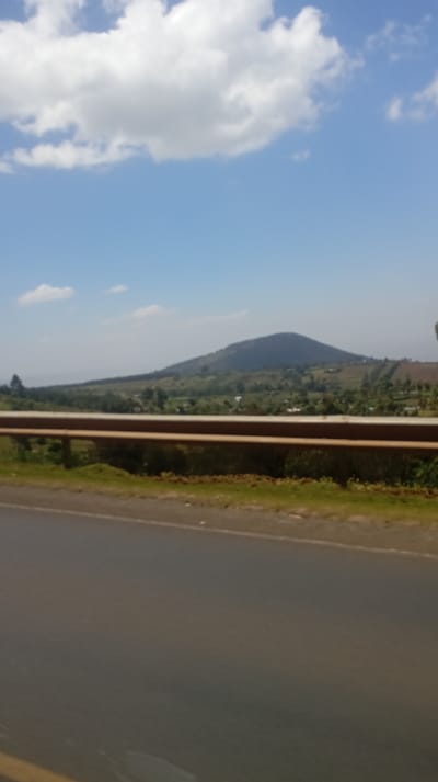 A road stretches into the distance under a blue sky dotted with white clouds. A rusted metal guardrail separates the road from a green landscape, where a conical mountain rises. The mountain is partially covered in trees and shrubs, with a few buildings nestled on its lower slopes. The overall scene depicts a rural or countryside environment.