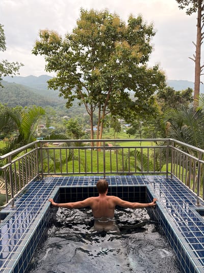 A man with blond hair is submerged in a dark blue tiled hot tub. The hot tub is outdoors, on a balcony or patio, overlooking a lush green landscape. The man is facing away from the camera, with his arms resting on the edge of the hot tub. The background consists of rolling hills covered in dense vegetation, with a few scattered buildings visible in the distance. Tall trees, including palm trees, frame the scene. The sky is overcast. The location is identified as Ban Kao Dua, Thailand.