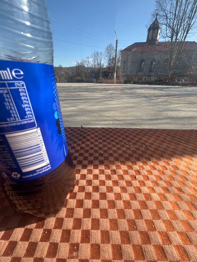 The scene is set in Brezovo, Bulgaria, under clear blue skies with direct sunlight. The foreground features a partially visible clear plastic bottle of Pepsi, angled slightly, with its dark brown liquid content and blue label prominently displayed. The label shows nutritional information in white text, a barcode, and Bulgarian text indicating "pepsi.bg" and "facebook.com/PepsiBulgaria". The bottle rests on a brown and light brown checkered textile surface, likely a tablecloth, exhibiting minor debris.
Beyond the immediate foreground, a wide, empty expanse of light grey asphalt pavement, resembling a public square or parking area, is visible. Long, distinct shadows are cast across this paved surface from an unseen source, indicating a low sun angle. In the mid-ground, a tall, grey utility pole with overhead power lines stands.
The background is dominated by a historical-style stone building with a red tiled roof and a multi-tiered bell tower, consistent with a church. The building features arched windows. To the right and behind the building, several large deciduous trees with bare branches are present. Distant hills or mountains can be seen on the horizon. There are no individuals present in the visible area.